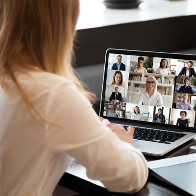 View over businesswoman shoulder sit at workplace desk looks at pc screen during group videocall with different age and ethnicity colleagues using application and webcam, distant communication concept