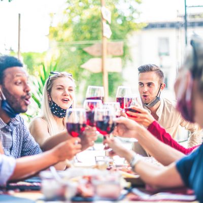 Friends toasting red wine at outdoor restaurant bar with face mask down - New normal lifestyle concept with happy people having fun together - Focus on guys behind