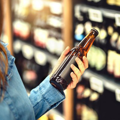 Customer buying beer in liquor store. Lager, craft or wheat beer. IPA or pale ale. Woman at alcohol shelf. Drink section and aisle in supermarket. Lady holding bottle in hand. Drink business concept.