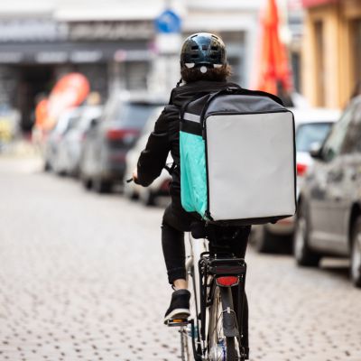 Rear view of a food delivery boy on a bike in city. Courier person riding bicycle with thermal backpack on city street for food delivery..