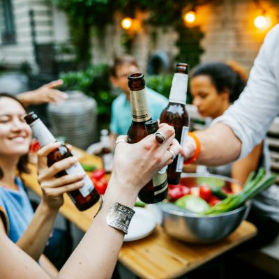 A group of friends clink beers as they make a toast at a barbecue meetup together.